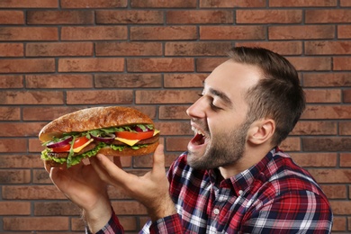 Photo of Young hungry man eating tasty sandwich on brick wall background