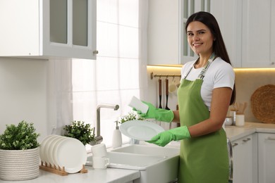 Photo of Happy woman in gloves washing plate above sink in kitchen, space for text