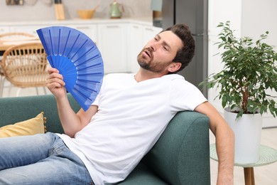Bearded man waving blue hand fan to cool himself on sofa at home