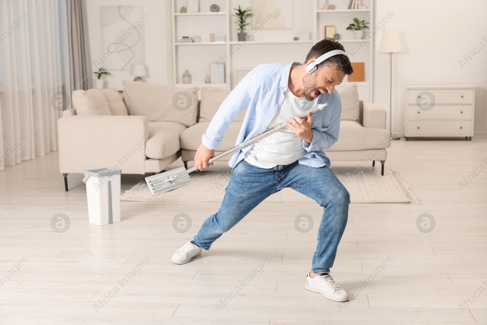 Photo of Enjoying cleaning. Happy man in headphones with mop singing while tidying up at home