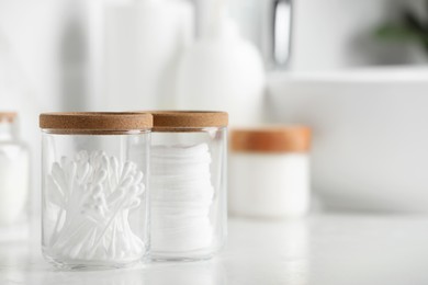 Glass jars with cotton pads and swabs on white countertop in bathroom, closeup. Space for text