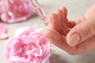 Photo of Woman dripping rose essential oil on finger against blurred flowers, closeup