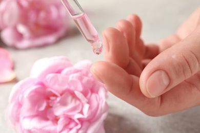 Woman dripping rose essential oil on finger against blurred flowers, closeup