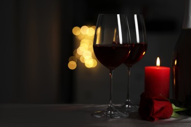 Photo of Glasses of red wine, rose flower and burning candle on grey table against blurred lights, space for text. Romantic atmosphere