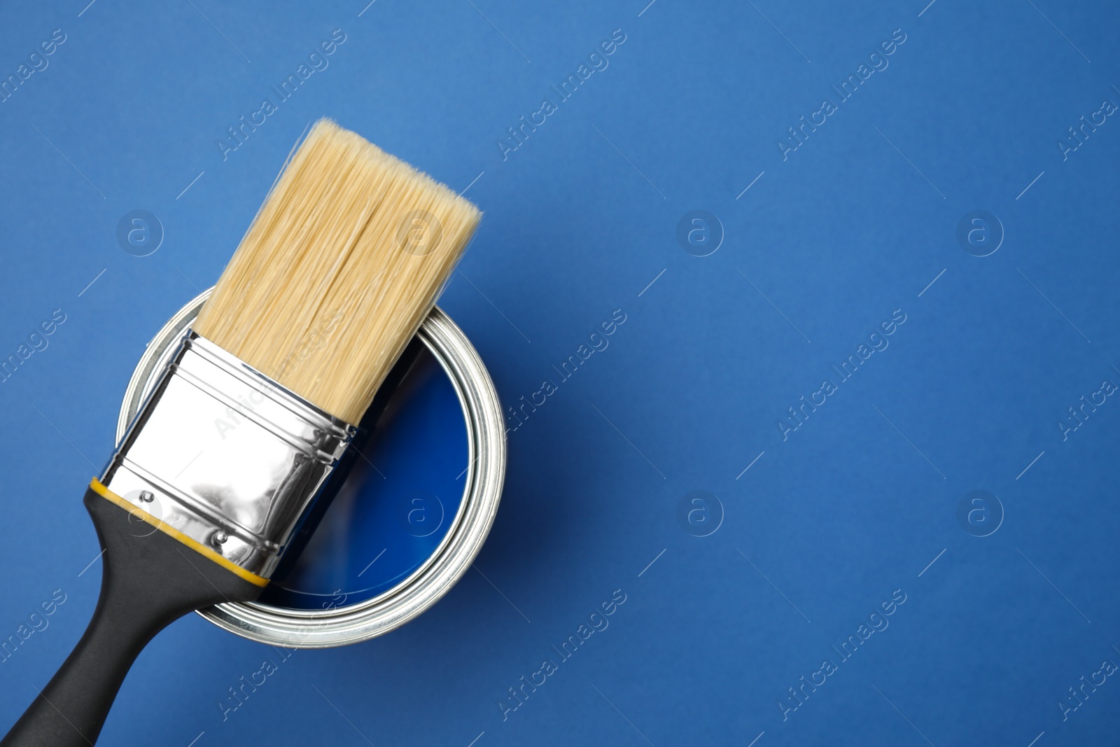 Photo of Open can with paint and brush on blue background, top view. Space for text