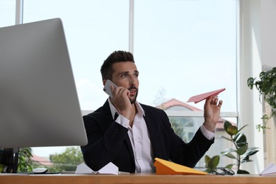 Photo of Handsome businessman playing with paper plane while talking on smartphone at desk in office
