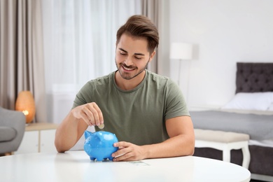 Photo of Young man putting coin into piggy bank at table indoors