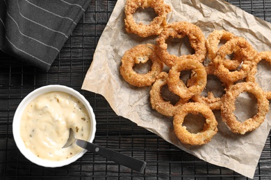 Photo of Cooling rack with homemade crunchy fried onion rings and sauce on wooden background, top view