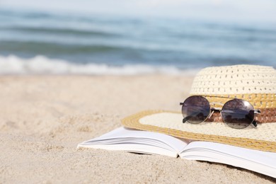 Photo of Open book, sunglasses and hat on sandy beach near sea, closeup. Space for text