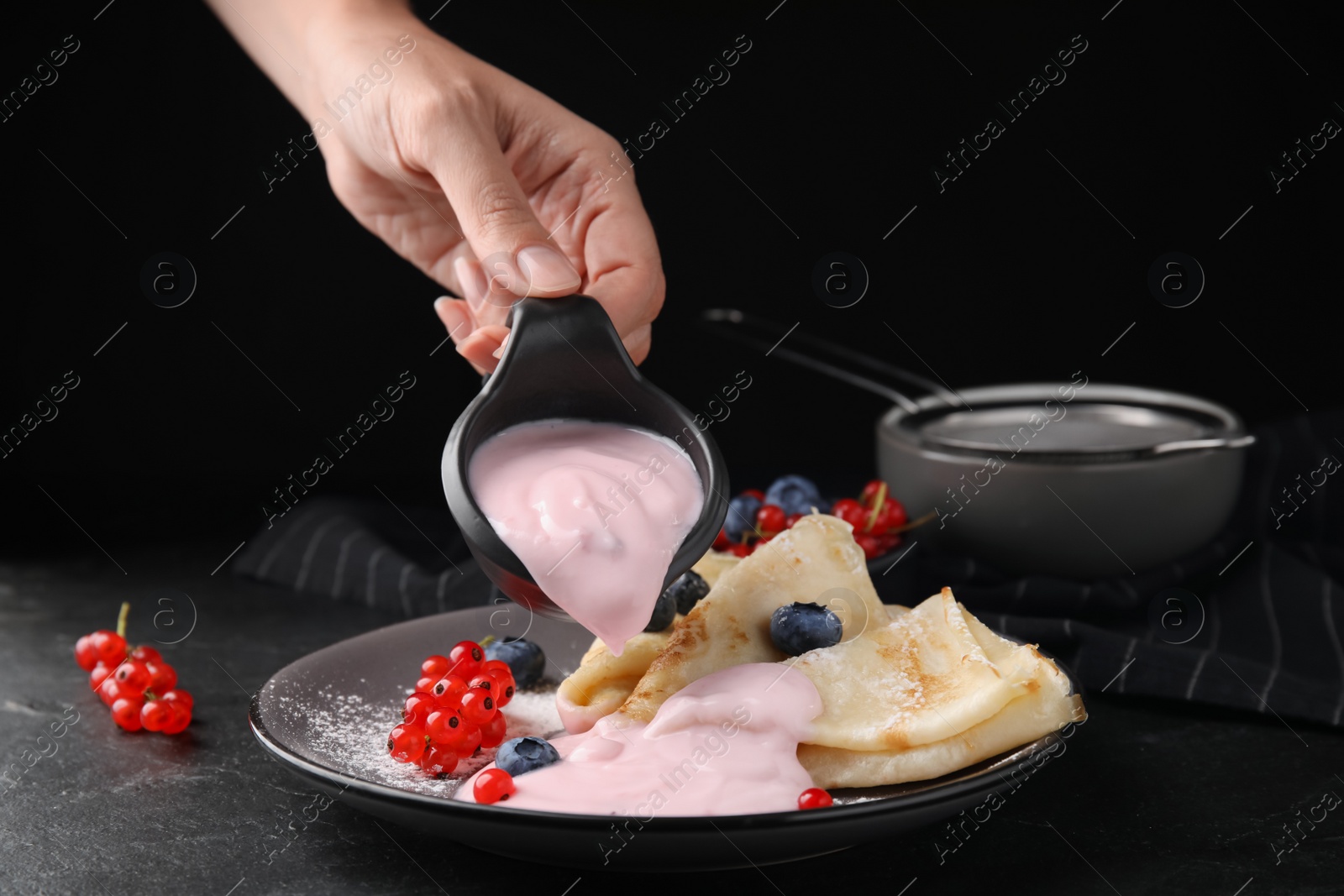 Photo of Woman pouring natural yogurt onto crepes with blueberries and red currants at table, closeup