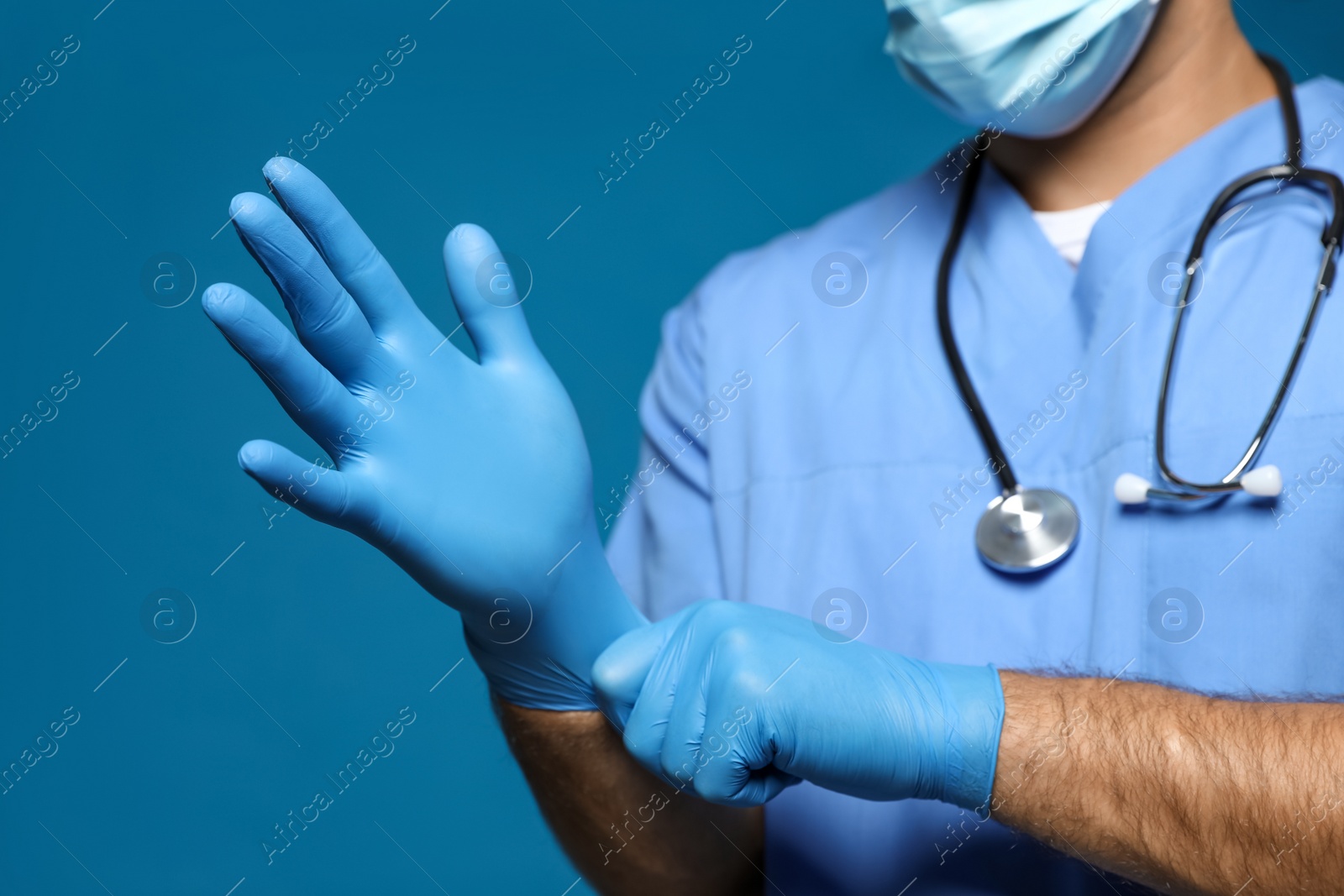 Photo of Doctor in protective mask putting on medical gloves against blue background, closeup