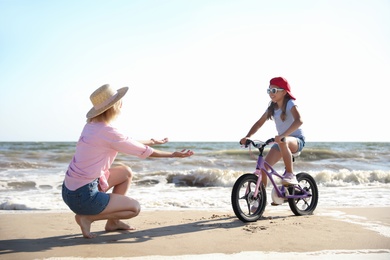 Happy mother teaching daughter to ride bicycle on sandy beach near sea