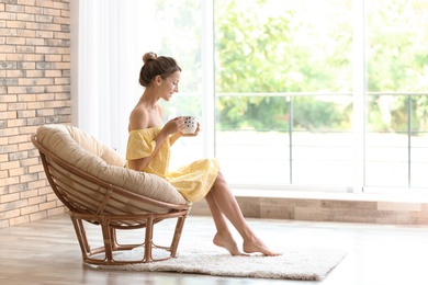 Photo of Young woman with cup of aromatic coffee sitting in papasan chair near window at home