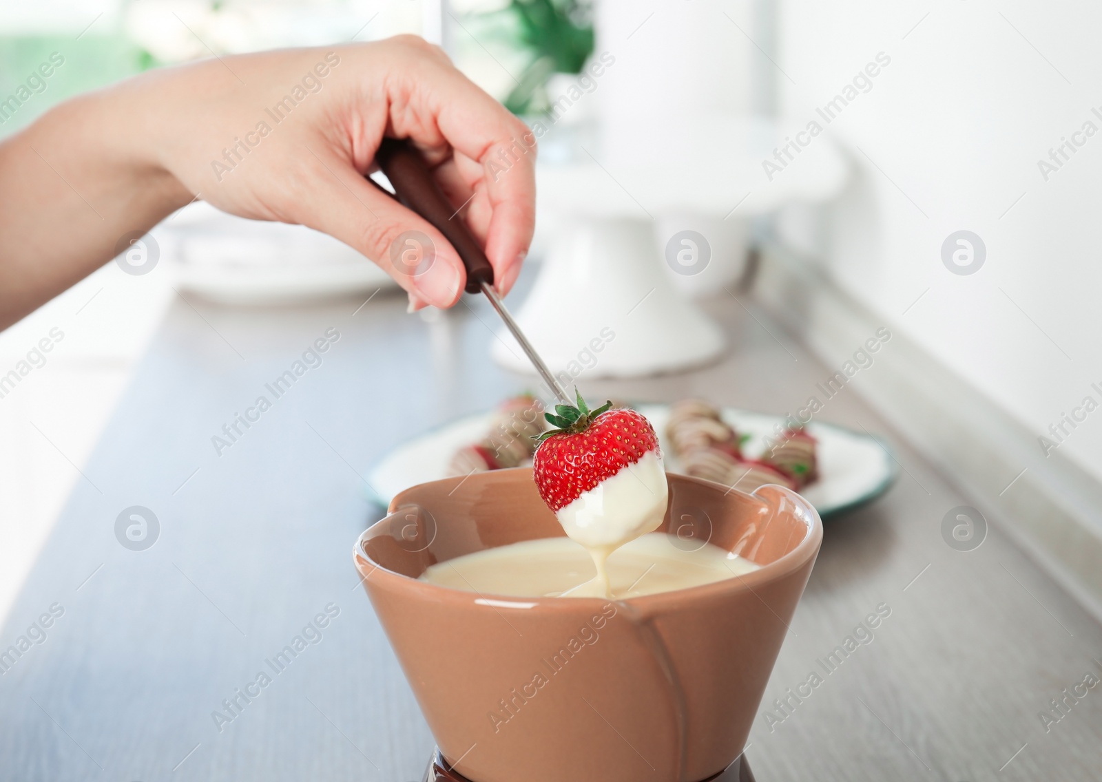 Photo of Woman dipping ripe strawberry into bowl with white chocolate fondue on table