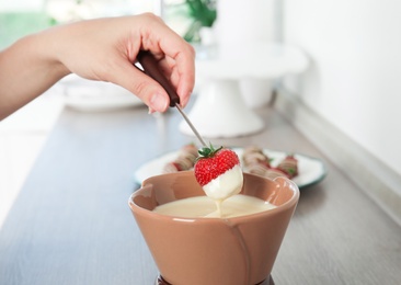 Woman dipping ripe strawberry into bowl with white chocolate fondue on table