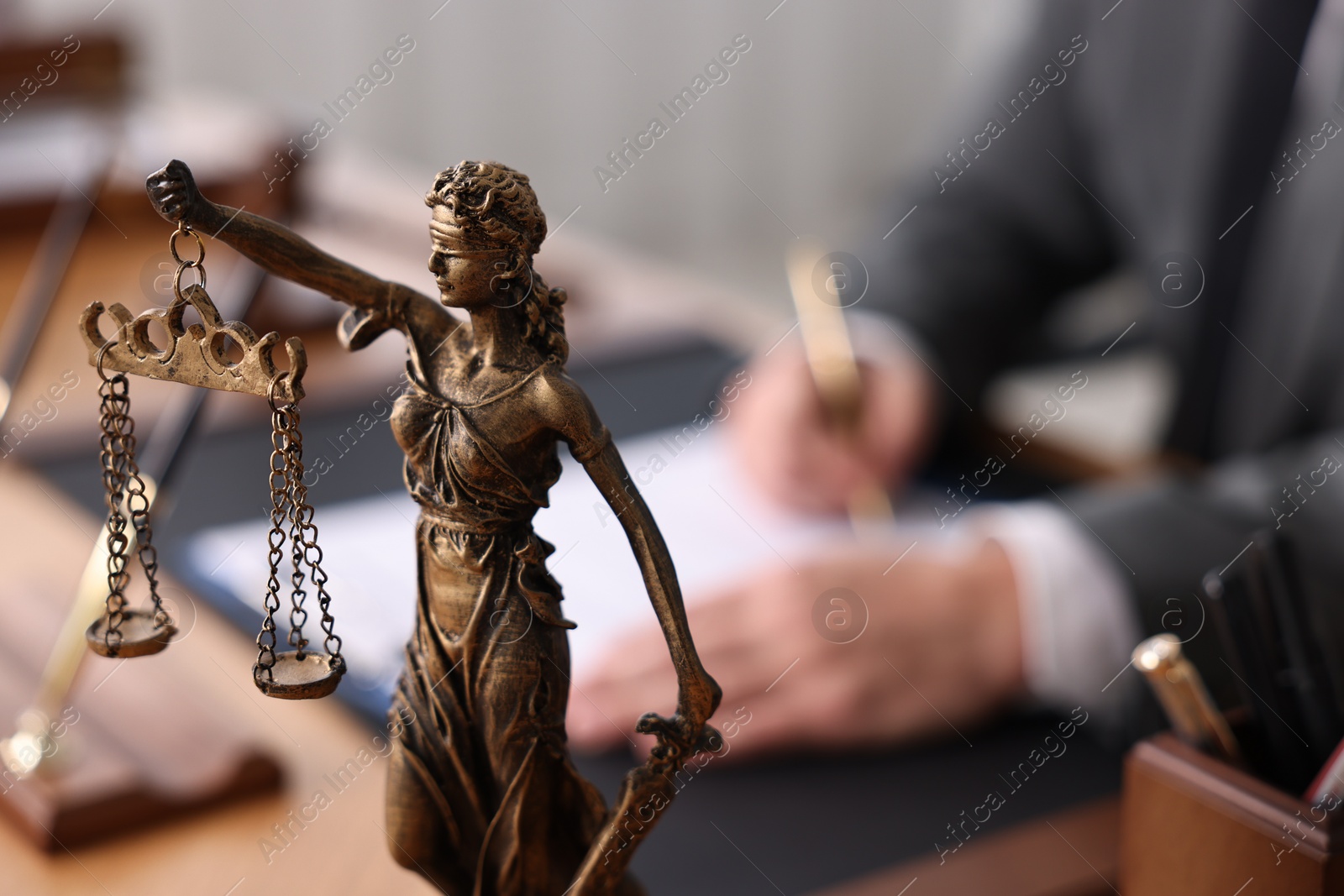 Photo of Notary writing notes at wooden table in office, focus on statue of Lady Justice