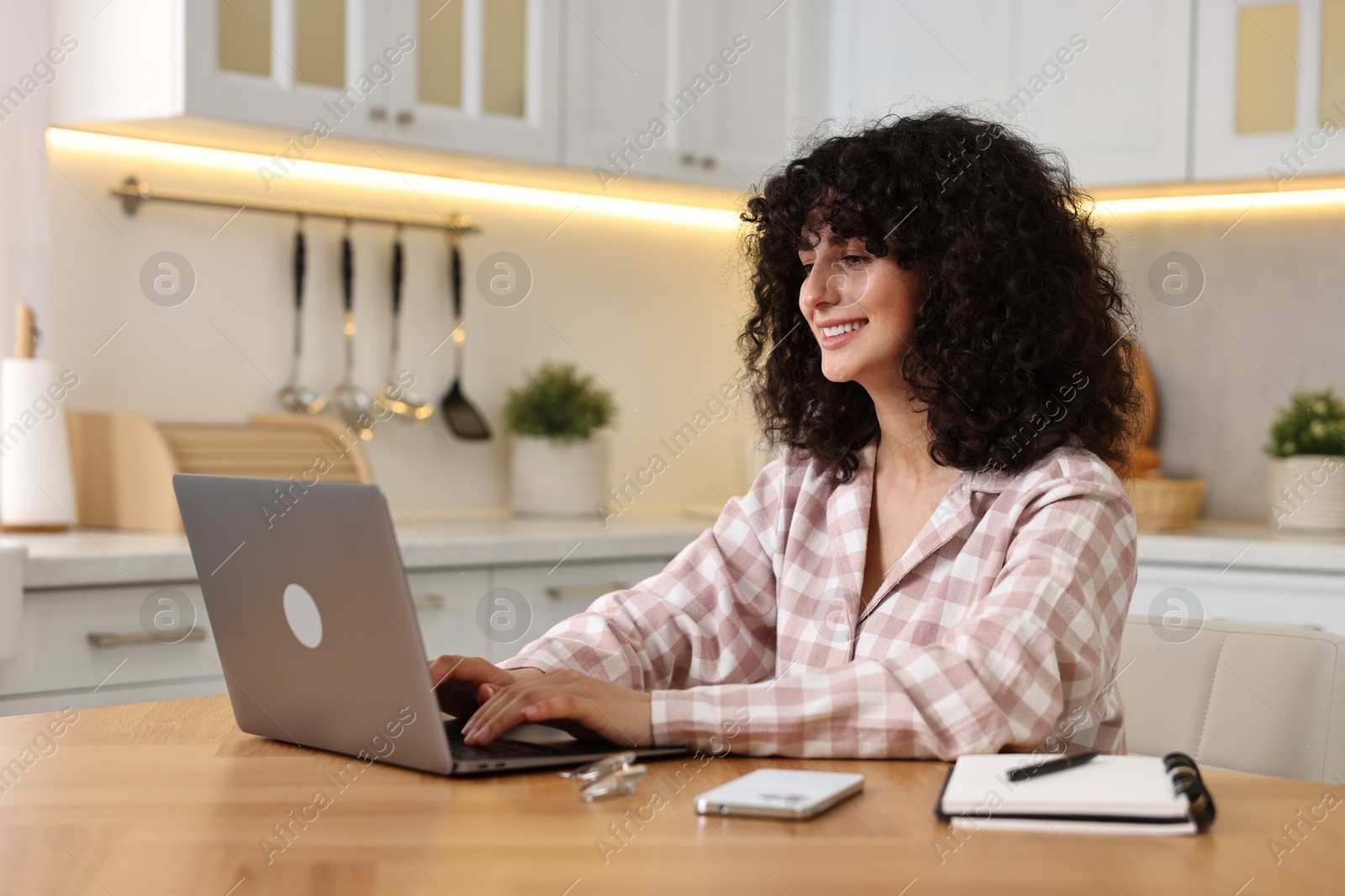 Photo of Beautiful young woman in stylish pyjama using laptop at wooden table in kitchen