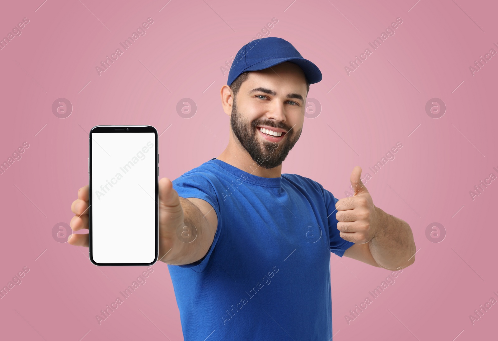 Image of Happy courier holding smartphone with empty screen and showing thumbs up on pink background