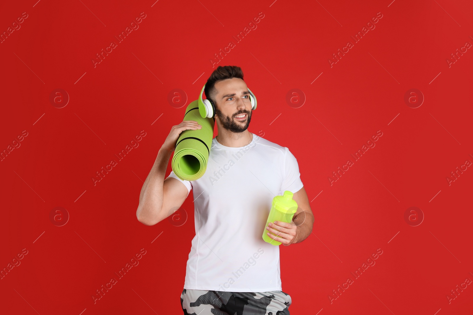 Photo of Handsome man with headphones,yoga mat and shaker on red background