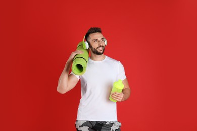 Handsome man with headphones,yoga mat and shaker on red background
