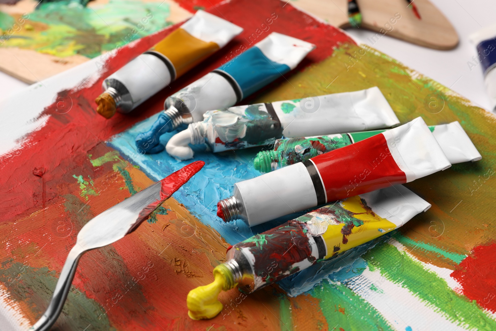 Photo of Tubes of colorful oil paints, spatula and canvas with abstract painting on table, closeup