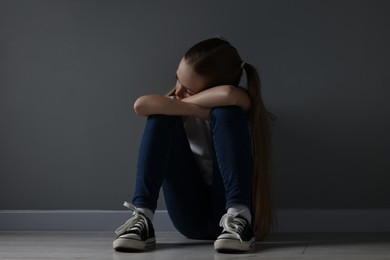 Sad girl sitting on floor near dark grey wall indoors