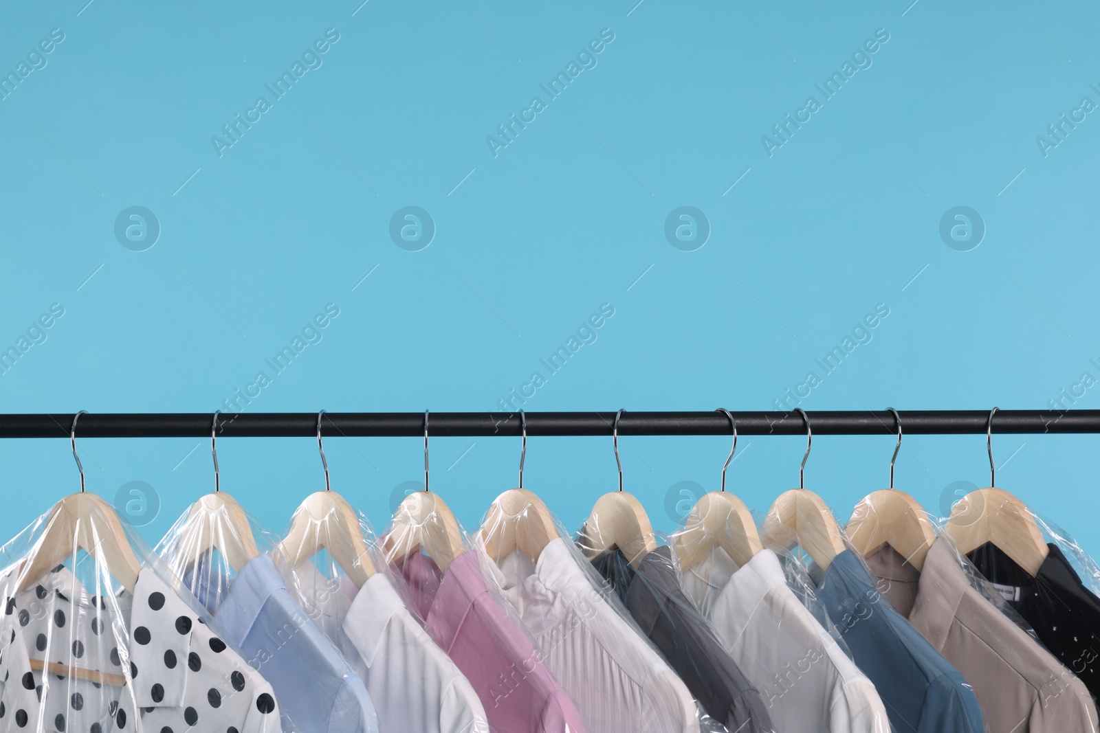 Photo of Dry-cleaning service. Many different clothes in plastic bags hanging on rack against light blue background, space for text