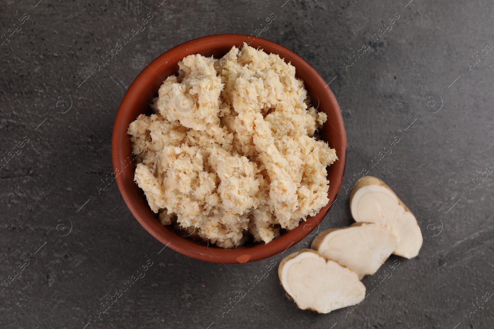 Photo of Bowl of tasty prepared horseradish and cut root on grey table, flat lay