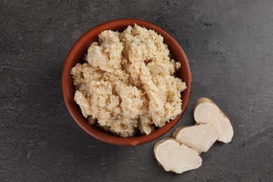 Bowl of tasty prepared horseradish and cut root on grey table, flat lay
