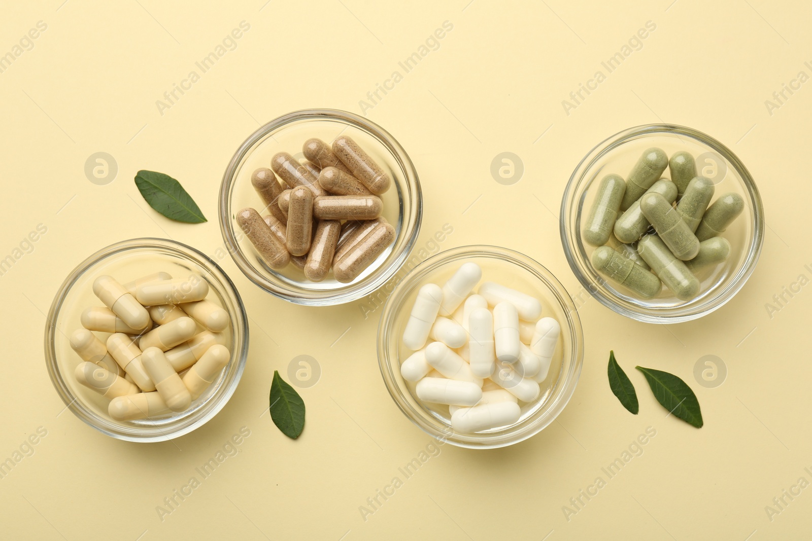 Photo of Different vitamin capsules in bowls and leaves on pale yellow background, flat lay