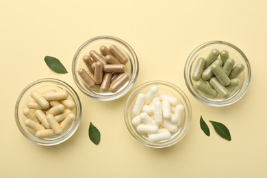 Photo of Different vitamin capsules in bowls and leaves on pale yellow background, flat lay