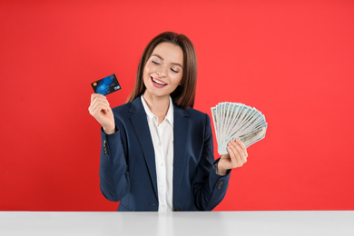 Young woman with money and credit card at table on crimson background