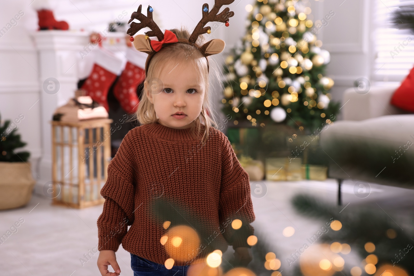 Photo of Cute little girl wearing deer headband in room decorated for Christmas