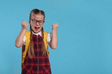 Emotional schoolgirl in glasses with backpack on light blue background, space for text