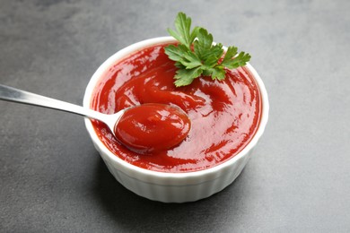 Photo of Taking delicious tomato ketchup with spoon from bowl at grey textured table, closeup