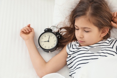 Photo of Cute little girl with alarm clock sleeping in bed, above view