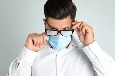 Photo of Man wiping foggy glasses caused by wearing medical mask on light background