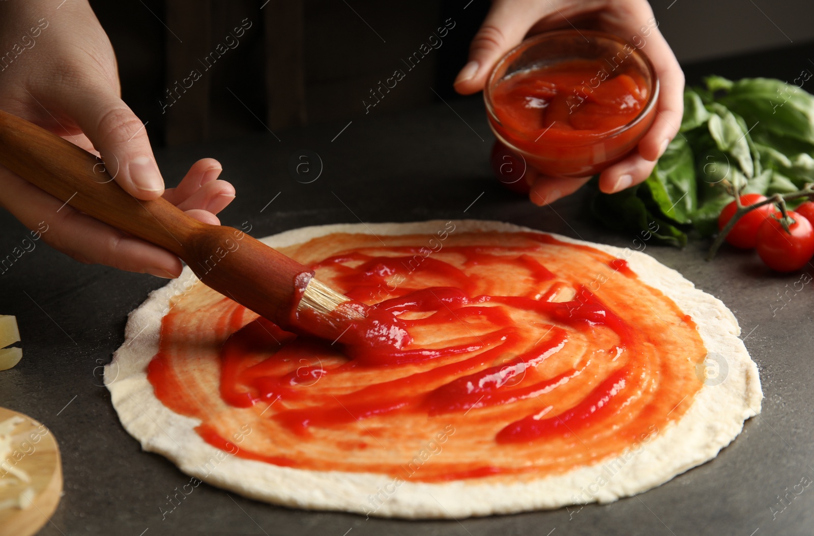 Photo of Woman spreading tomato sauce onto pizza crust at grey table, closeup