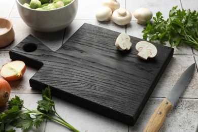 Photo of Black wooden cutting board and products on light tiled table, closeup