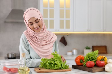 Muslim woman making delicious salad with vegetables at white table in kitchen. Space for text