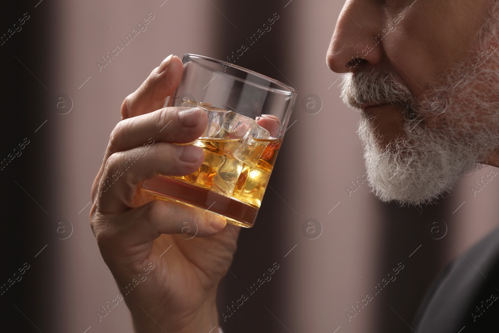 Photo of Senior man holding glass of whiskey with ice cubes on brown background, closeup