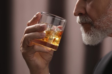 Photo of Senior man holding glass of whiskey with ice cubes on brown background, closeup