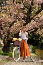 Beautiful young woman with bicycle and flowers in park on pleasant spring day