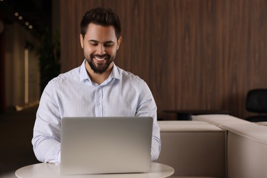 Happy young man working on laptop at table in office. Space for text