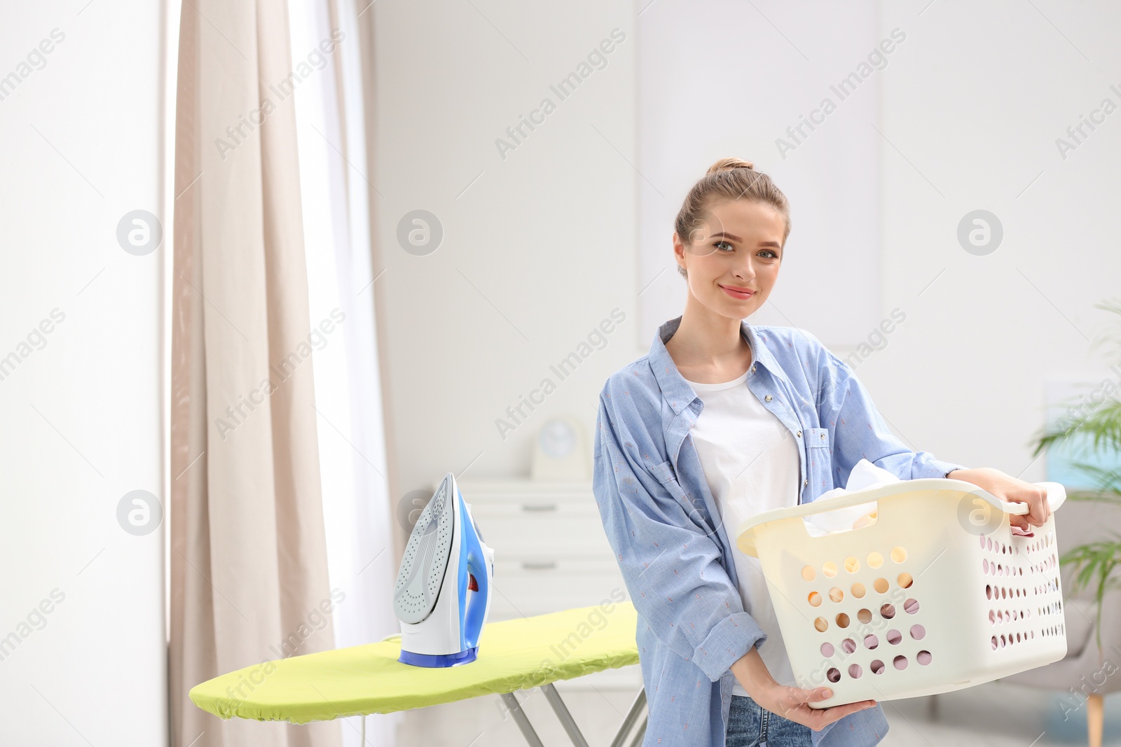 Photo of Young pretty woman holding basket of clean laundry at ironing board indoors. Space for text