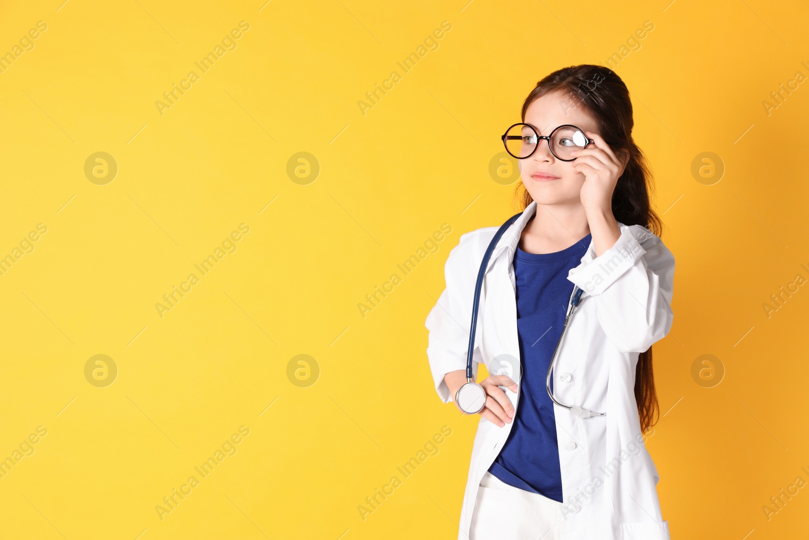 Photo of Little girl with eyeglasses and stethoscope dressed as doctor on yellow background, space for text. Pediatrician practice