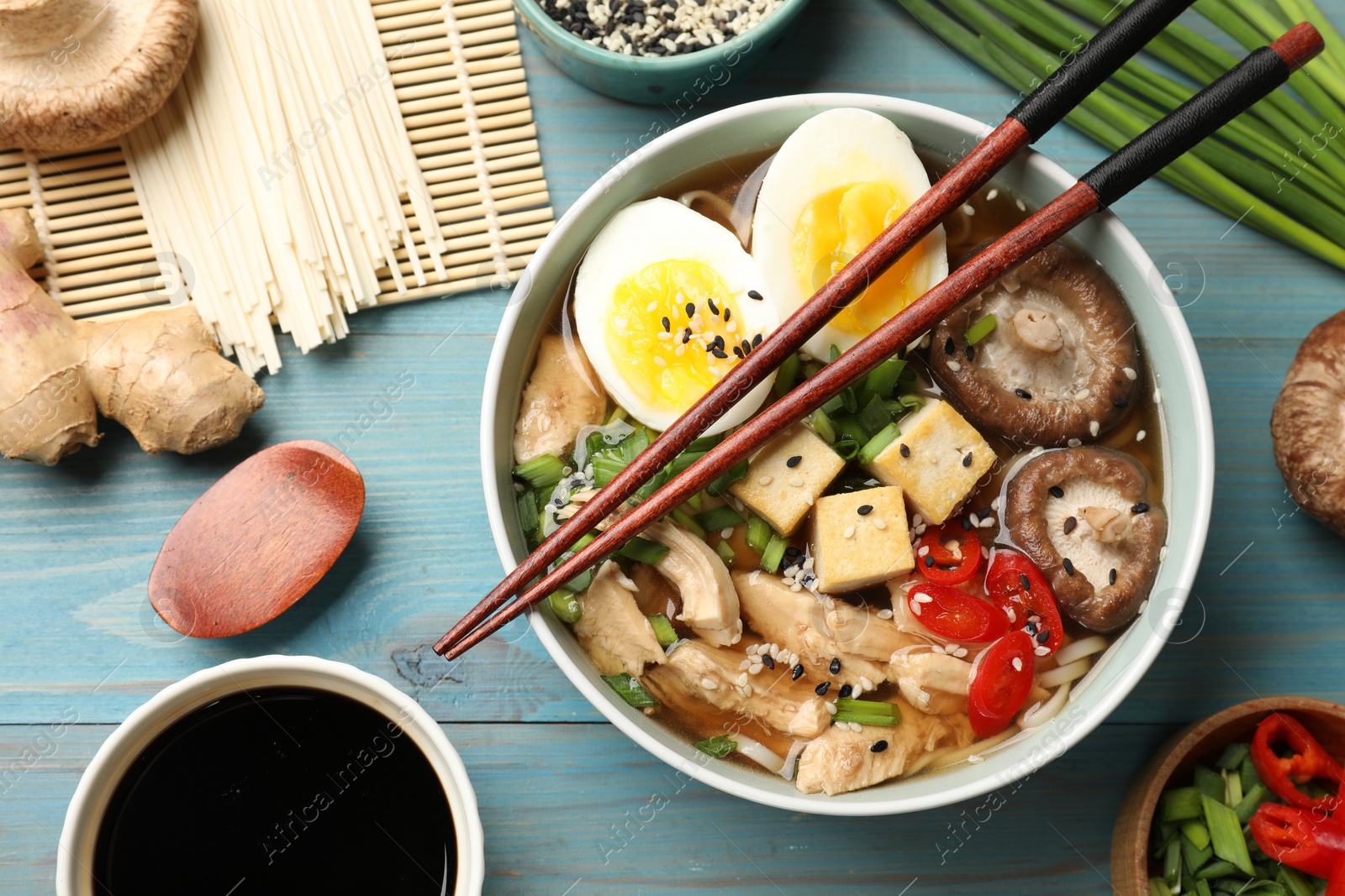 Photo of Bowl of delicious ramen and ingredients on light blue wooden table, flat lay. Noodle soup