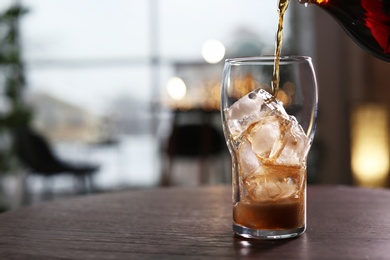 Photo of Pouring cola into glass with ice on table against blurred background. Space for text