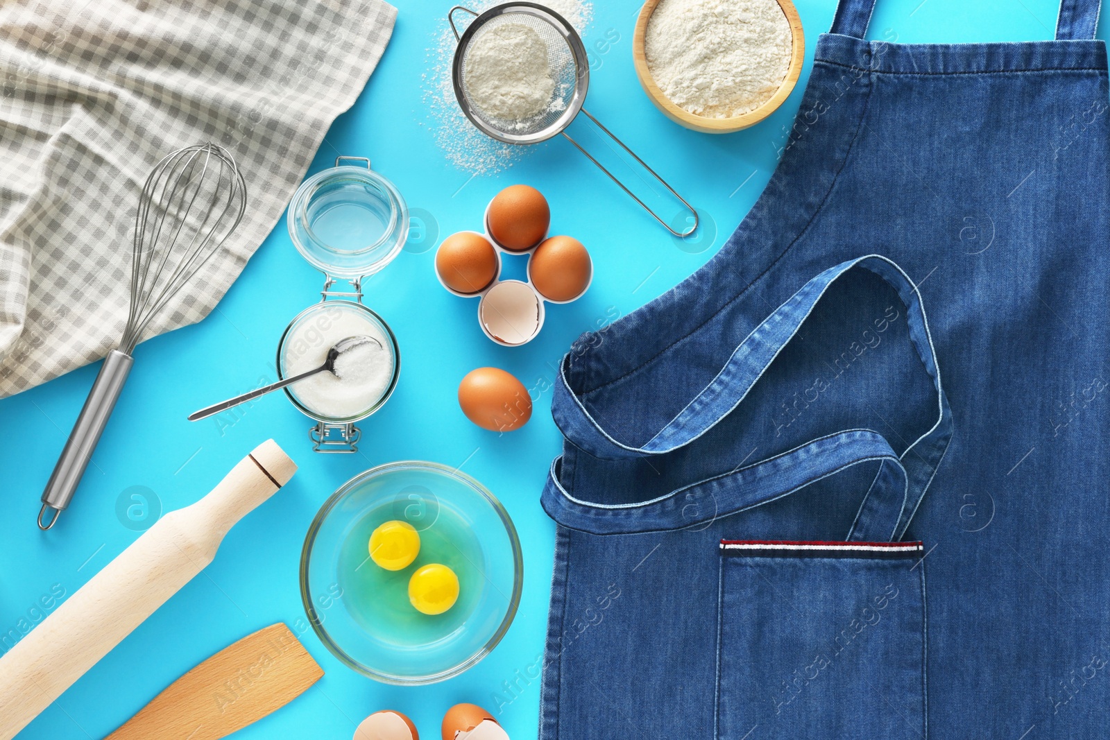 Photo of Denim apron, different ingredients and kitchen tools on light blue background, flat lay