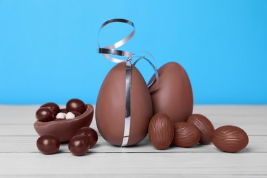Delicious chocolate eggs and candies on white wooden table against light blue background, closeup
